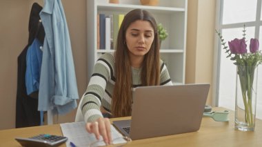 Odaklanmış İspanyol bir kadın evde dizüstü bilgisayarı, hesap makinesi ve dokümanlarıyla çalışıyor..