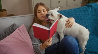 Oturma odasındaki kanepede kitap okuyan genç sarışın bir kadın yanında beyaz bir köpekle..
