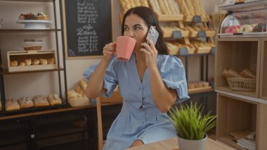 Genç esmer kadın kahve içiyor ve bir fırında telefonla konuşuyor. Arkasında ekmek ve hamur işleri rafları var.