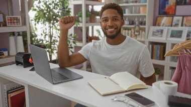 Ev dekorunda gülümseyen genç bir adam dizüstü bilgisayarı ve masasında bir not defteriyle.