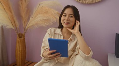 Genç bir kadın, arka planda yumuşak mor bir duvarla rahat bir oturma odasında tablet okurken gülümsüyor..
