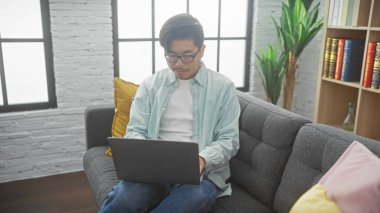 Asyalı adam modern bir oturma odasında dizüstü bilgisayar kullanıyor tuğla duvar ve yeşil bitkilerle.
