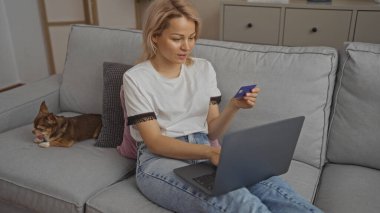 Sarışın, beyaz bir kadın oturma odasında elinde bir çuvava, elinde bir kredi kartı ve dizüstü bilgisayar ile oturuyor..