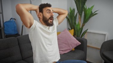 Kapalı bir koltukta oturan sakallı, şaşırmış bir adam. Oturma odasında elleri arkada şokunu ifade ediyor..