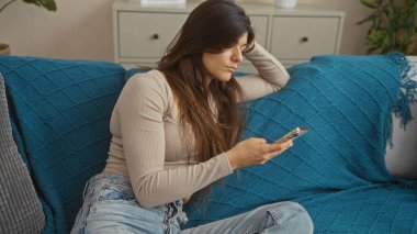 Esmer saçlı genç bir İspanyol kadın evdeki mavi kanepesinde dinlenirken telefonunu kullanıyor..