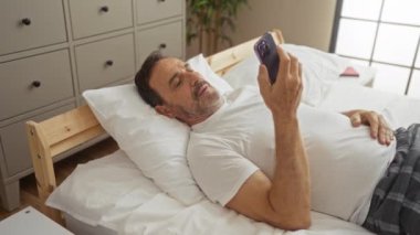 Yatak odasında orta yaşlı bir adam şaşırmış bir şekilde akıllı telefon kullanıyor, kapalı bir evde yatakta yatıyor.