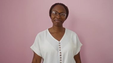 Genç Afro-Amerikan kadın gözlük takıyor, mutlu bir jestle duruyor, tamam parmaklarından bakıyor, pembe arka planda sırıtıyor.