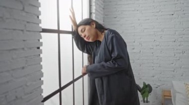 Modern bir yatak odasının penceresine yaslanmış, tuğla duvarlı arka planı olan sıkıntılı bir kadın.