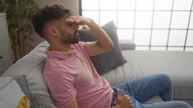 Pembe gömlekli, sakallı genç adam, modern dekorlu, parlak oturma odasında, eli alnında kanepede dinleniyor.