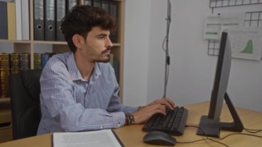 Evraklar ve klasörlerle çevrili bir bilgisayarda çalışan genç adam profesyonel bir iç mekanda çalışıyor..