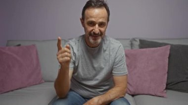 Bir evde oturan İspanyol bir adam, gri bir kanepede gri yastıklı günlük gri gömlek ve mavi kot pantolon giyerek, düşünceli bir ifadeyle başını kaldırıp dokunuyor.