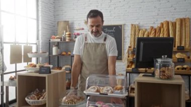 Ekmek rafları, glütensiz tabelalar ve yazar kasayla çevrili çeşitli hamur işleri sergilenen bir pastanede çalışan önlüklü orta yaşlı bir adam.
