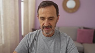 Beyaz saçlı, sakallı, gri tişörtlü, mor duvarlı ve arka planda yuvarlak aynalı İspanyol bir adam oturma odasında oturuyor.