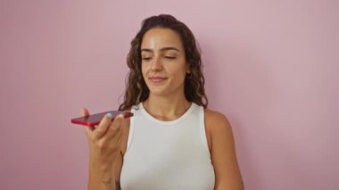 İspanyol kadın telefonundan sesli mesaj yolluyor, beyaz bir atlet giyerken pembe bir arka plana karşı duruyor..