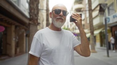 Beyaz saçlı, güneş gözlüklü ve sakallı bir adam elinde akıllı bir telefonla şehir sokağında dikiliyor.