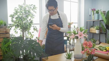 Genç Asyalı bir adam yemyeşil çiçeklerle dolu canlı bir çiçekçide çalışırken akıllı telefonunu kontrol ediyor..