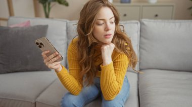 Sarı kazaklı genç sarışın bir kadın oturma odasındaki açık gri bir kanepede, elinde akıllı bir telefonla, rahat bir evde düşünceli ve çekici görünüyor..