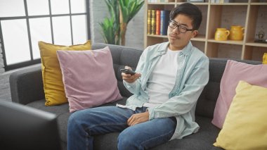 Gözlüklü genç bir Asyalı evde gri bir koltukta oturuyor, elinde sıkılmış bir ifadeyle uzaktan kumanda tutuyor..