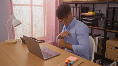 Kapalı ofis ortamında dizüstü bilgisayarı ve arka planda kitaplığı olan yakışıklı İspanyol bir adam..