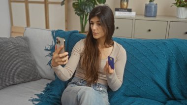 Oturma odasındaki koltukta oturan genç bir İspanyol kadın elinde akıllı bir telefon ve kredi kartıyla dikkatle telefona bakıyor.