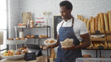 Fırında elinde hamur işleriyle gezen genç bir adam önlük takıyor. Etrafı taze ekmek ve tatlılarla çevrili. İyi aydınlatılmış bir dükkan ortamında sergileniyor.