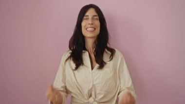 Neşeyle kutlayan bir kadın, uzun siyah saçlı çekici bir yetişkin, bej bir gömlek giyiyor, izole edilmiş pembe bir arka planda heyecan ve mutluluğu ifade ediyor.