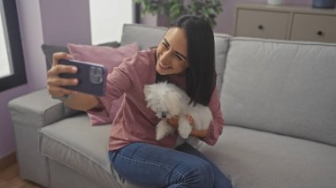 Gülümseyen kadın rahat bir oturma odasındaki kanepede pofuduk köpeğiyle selfie çekiyor..