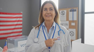 Mature woman doctor pointing at her 'i voted' sticker indoors with american flag and ballot boxes suggesting civic duty. clipart