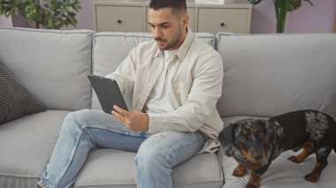 Genç İspanyol bir adam, modern bir oturma odasındaki kanepede, tekel bir köpeğin yanında rastgele bir tablet kullanmış..
