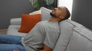 Modern oturma odasındaki kanepede dinlenen genç adam rahat bir ifadeyle, rahat ve sakin bir ev atmosferiyle şık bir dekor ve doğal ışıklandırmayla..