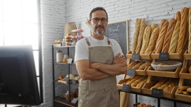 Gözlüklü ve önlüklü bir İspanyol fırında kolları çapraz duran çeşitli ekmeklerle çevrili bir adam.