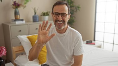 Orta yaşlı İspanyol bir adam yatağın yanındaki şifoniyerde bitkiler ve kitaplarla parlak bir yatak odasında el sallıyor.