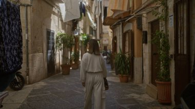 Genç İspanyol bir kadın asılmış giysiler ve yeşilliklerle çevrili, Bari, İtalya 'nın büyüleyici eski sokaklarında yürüyor, Puglia' nın tarihi güzelliğini yakalıyor..