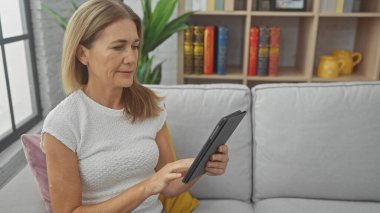 Modern oturma odasındaki koltukta tablet kullanan olgun bir kadın.