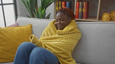 Genç, Afro-Amerikan bir kadın, modern bir oturma odasında, rahat sarı bir battaniyeye sarılmış bir kanepede dinleniyor..