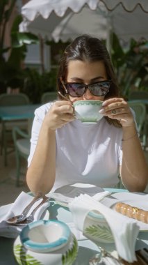 Güneş gözlüklü İspanyol kadın lüks bir terasta kahve yudumluyor. Etrafı şık sofra takımlarıyla çevrili. Açık havada sakin bir restoran deneyimini vurguluyor..