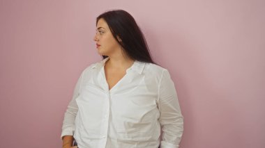 Pembe bir arka planda duran kıvrımlı vücutlu güzel genç bir kadın beyaz bir gömleğe olan güvenini gösteriyor.