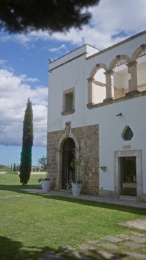 Puglia, İtalya 'da tarihi bir binanın çarpıcı dış manzarası, kırsal bir taş cephesi, yemyeşil çimenler, uzun selvi ağaçları ve dağınık bulutlu güzel mavi bir gökyüzü..