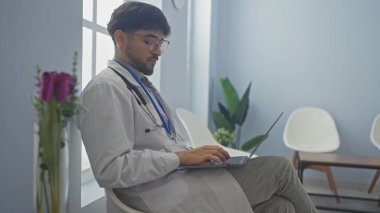 Tıbbi kıyafetli genç sakallı bir adam elinde dizüstü bilgisayarla parlak bir hastane lobisinde oturuyor..