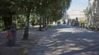 Roma 'daki villa borghese bahçelerinin bulanık manzarası. Ağaçların ve gölgelerin arasında yürüyen insanlar. Huzurlu, rahat bir yeşillik ve güneş ışığı atmosferi yaratıyor..