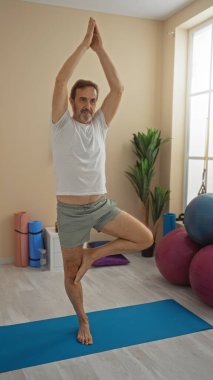 Sağlık merkezinde yoga yapan bir adam egzersiz ekipmanları ve sağlıklı yaşam tarzı vurgusu için canlı iç dekorasyon arasında denge ve konsantrasyon gösteriyor..