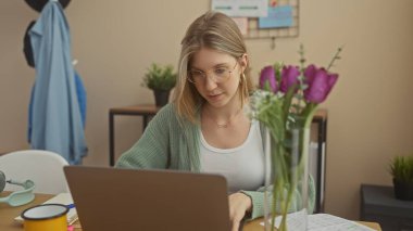 Modern bir ofiste dizüstü bilgisayarda çalışan sarışın bir kadın. Çiçekleri, gözlükleri ve rahat bir süveteri var.
