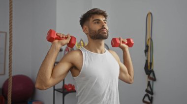 Yakışıklı bir adam spor salonunda dambıllarıyla egzersiz yapıyor, beyaz kolsuz bir gömlek giyiyor, spor salonuna odaklanıyor spor aletleriyle çevrili kararlı bir ifadeyle.