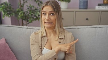 Oturma odasındaki genç, güzel, sarışın bir kadın evde rahat bir koltukta solunu göstererek kafa karışıklığını gösteren komik bir ifade takınıyor..