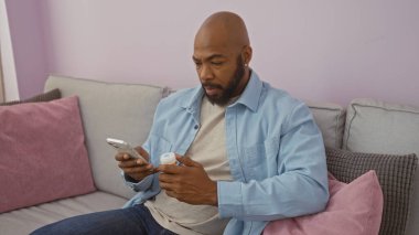 Genç Afro-Amerikalı bir adam, elinde ilaç şişesi ve akıllı telefonuyla apartman dairesinde oturuyor.