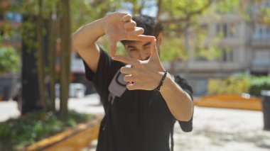 Genç bir adam, bir şehir parkında elleri ile kamerayı çerçeveler, yaratıcılık ve şehir yaşamını gösterir..