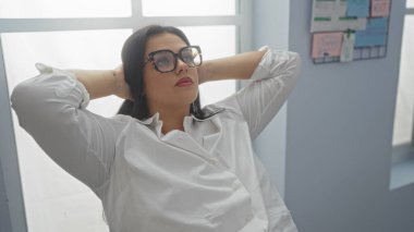 Ofis odasında gözlüklü, beyaz gömlekli, rahat genç bir kadın, arkasını yaslarken kendinden emin ve sakin görünüyor, profesyonel bir ortamda rahat ve sakin bir tavır sergiliyor.