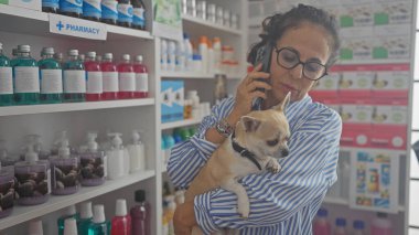 Orta yaşlı bir kadın renkli ürünlerle dolu bir eczanede telefonda bir chihuahua tutuyor..