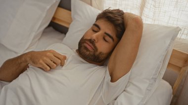 Yakışıklı genç adam güneşli bir yatak odasında uzanıyor, huzurlu ve rahat görünüyor, beyaz gömlekli ve rahat yastıklı.