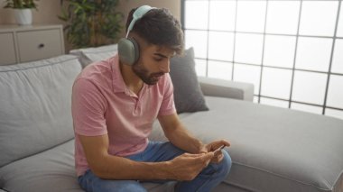 Sakallı, kulaklık takan, pembe gömlekli, modern bir oturma odasında oturan İspanyol bir adam. Telefonunu kullanıyor.